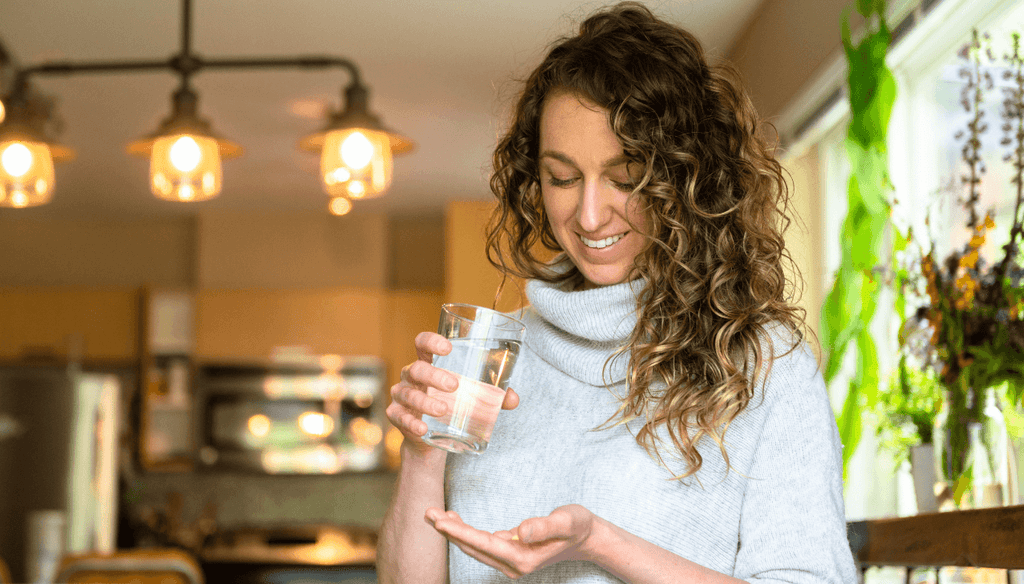 A happy woman at her house taking her supplements with a glass of water in her hand.
