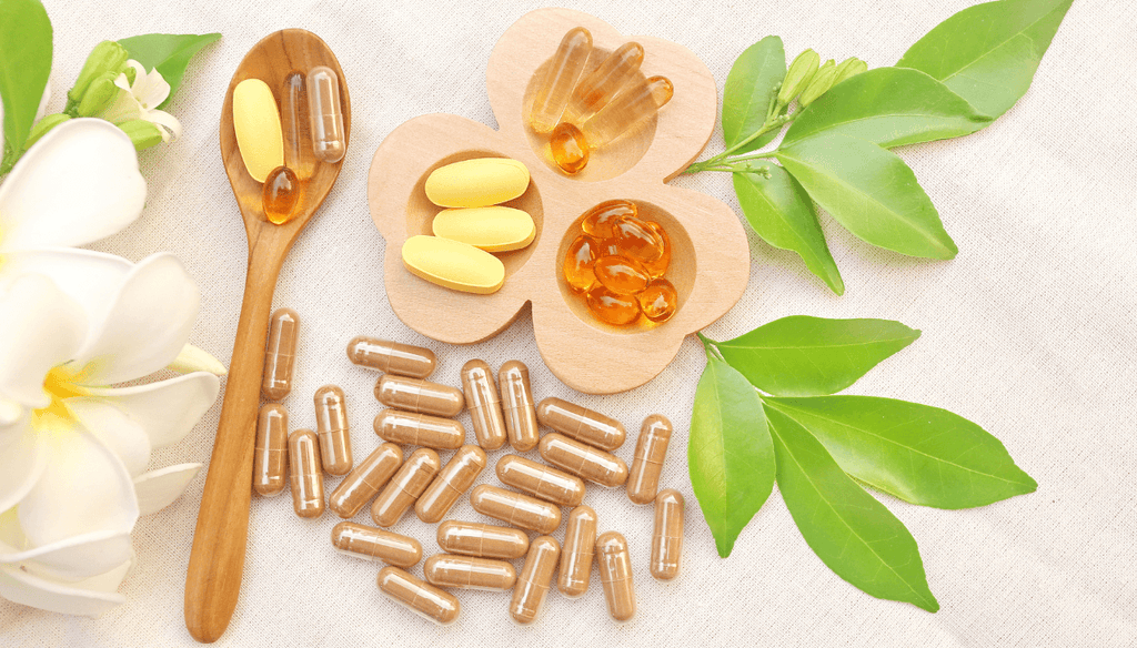 Different supplements and vitamins on a wood spoon and on the table plus some green leaves and white flower