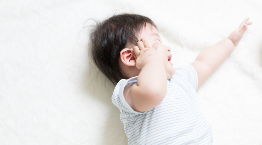 Baby lying on a bed on its side with its hand rubbing its ear and crying