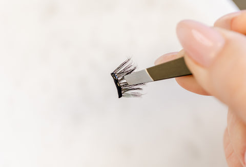 A picture of a white background a manicured hand holding tweezers that have a segment of eyelash extensions pinched between them. 