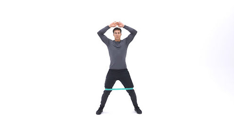 A man standing with his feet apart and his hands above his head with a resistance band around his knees