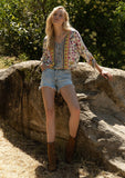 [Color: Natural/Yellow] A front facing image of a blonde model sitting outside wearing a classic bohemian summer top in a mixed floral print. With three quarter length sleeves, a gathered sleeve detail with ties, a v neckline, a self covered button front, and an elastic waist.