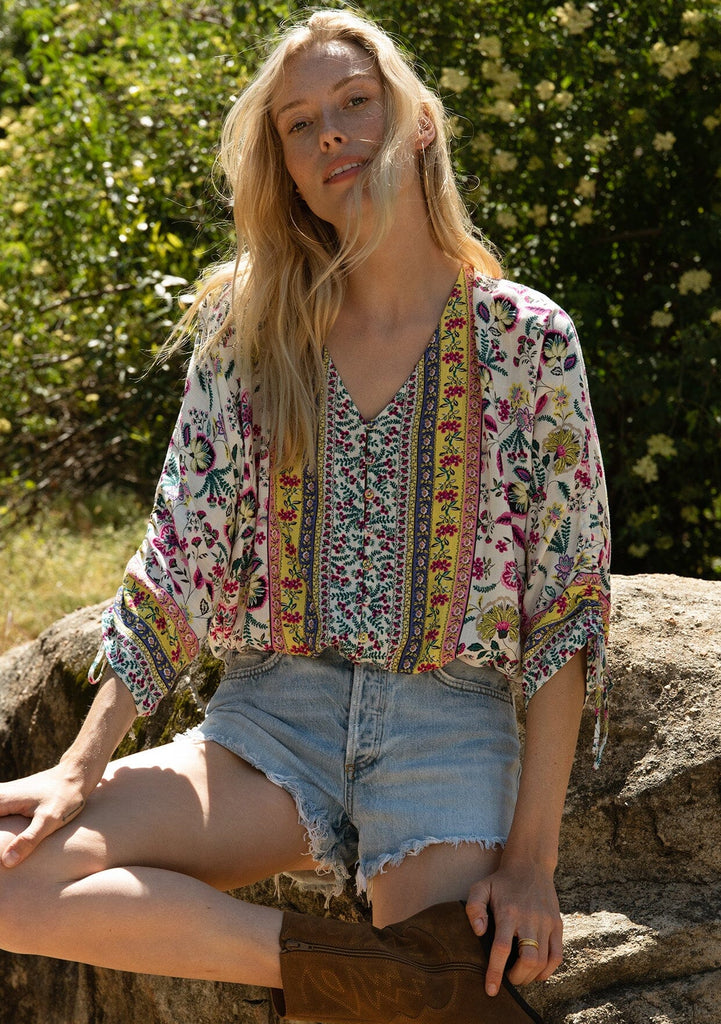 [Color: Natural/Yellow] A front facing image of a blonde model sitting outside wearing a classic bohemian summer top in a mixed floral print. With three quarter length sleeves, a gathered sleeve detail with ties, a v neckline, a self covered button front, and an elastic waist.