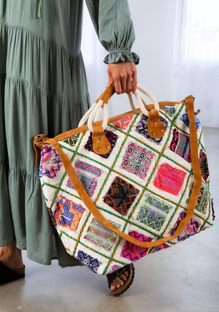 [Color: Multi/Natural] A model holding a patchwork weekender bag. With embroidered details, suede trim, suede top handles, and a suede long adjustable strap.