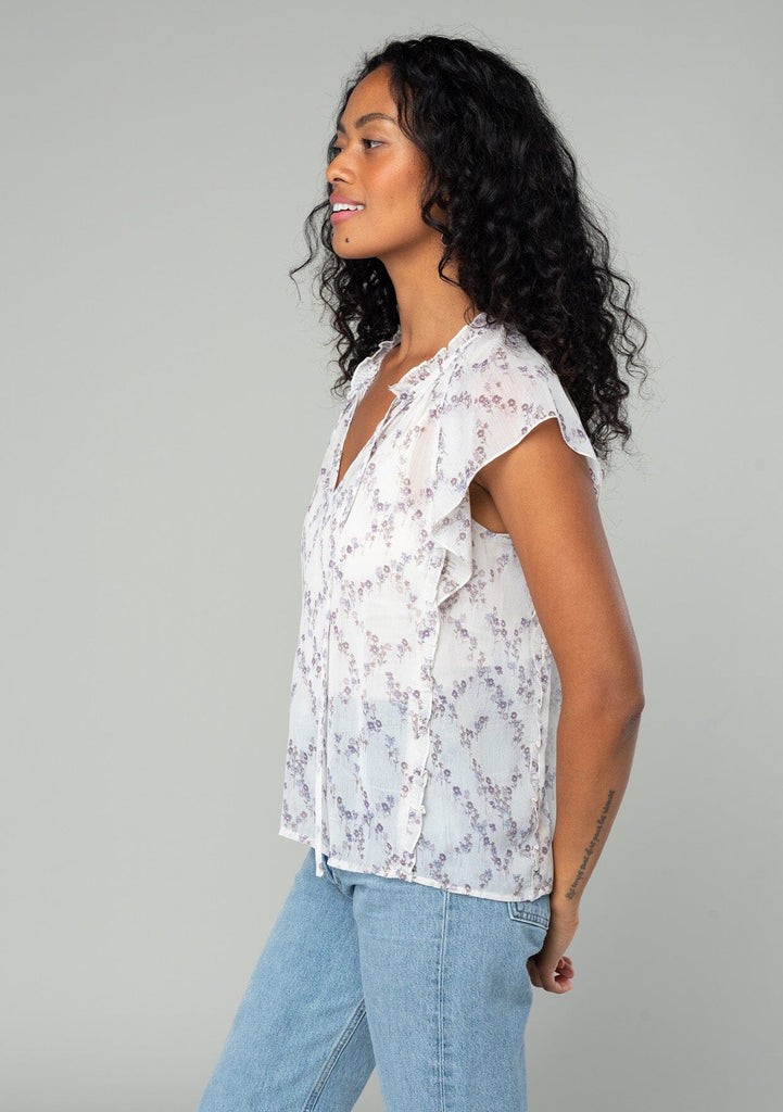 [Color: Ivory/Plum] A side facing image of a brunette model wearing a sheer chiffon bohemian spring top in an ivory white and plum purple floral print. With short flutter sleeves, a split v neckline with ties, and ruffled trim throughout. 