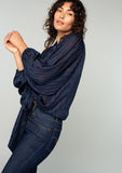 [Color: Navy] A side facing image of a brunette model wearing a sheer navy blue bohemian tie front top in a sparkly lurex stripe. With voluminous long sleeves and a tie front waist that can be styled in multiple ways. 