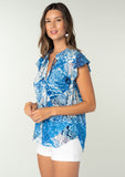 [Color: Blue/Navy] A side facing image of a brunette model wearing a bright blue floral patchwork print bohemian top. With a button front, short flutter sleeves, and neck ties. 