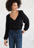 [Color: Black] A model wearing a Holiday ready, classic black peasant top in textured crepe, with beaded trim along the neckline. With a split neckline and tassel ties. A timeless blouse for your next special occasion. 