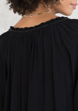 [Color: Black] A model wearing a Holiday ready, classic black peasant top in textured crepe, with beaded trim along the neckline. With a split neckline and tassel ties. A timeless blouse for your next special occasion. 