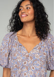 [Color: Grey/Natural] A close up front facing image of a brunette model wearing a classic bohemian blouse in a grey and purple mixed floral print. With short puff sleeves, a loop button front, and a split v neckline with tassel ties. 