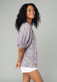 [Color: Grey/Natural] A side facing image of a brunette model wearing a classic bohemian blouse in a grey and purple mixed floral print. With short puff sleeves, a loop button front, and a split v neckline with tassel ties. 