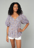 [Color: Grey/Natural] A half body front facing image of a brunette model wearing a classic bohemian blouse in a grey and purple mixed floral print. With short puff sleeves, a loop button front, and a split v neckline with tassel ties. 