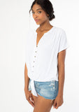 [Color: Chalk] A side facing image of a black model wearing a soft and silky white short sleeve top with a button front and tie waist detail. 