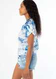 [Color: Navy/Teal] A side facing image of a black model wearing a blue and white watercolor floral print short sleeve top with a button front and a tie front detail at the waist. 