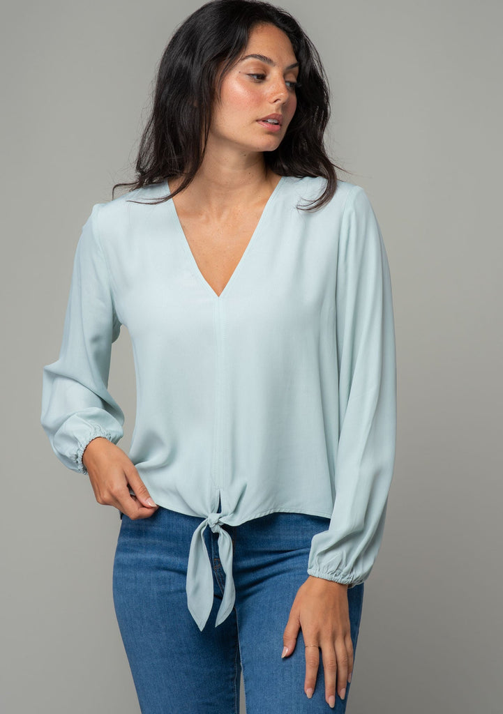 [Color: Sky] A front facing image of a brunette model wearing a soft and silky light blue crepe long sleeve top with a tie front detail. 