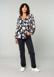 [Color: Black/Natural] A full body front facing image of a brunette model wearing a black and off white floral print blouse. A bohemian resort top with long sleeves and a wide elastic neckline. 