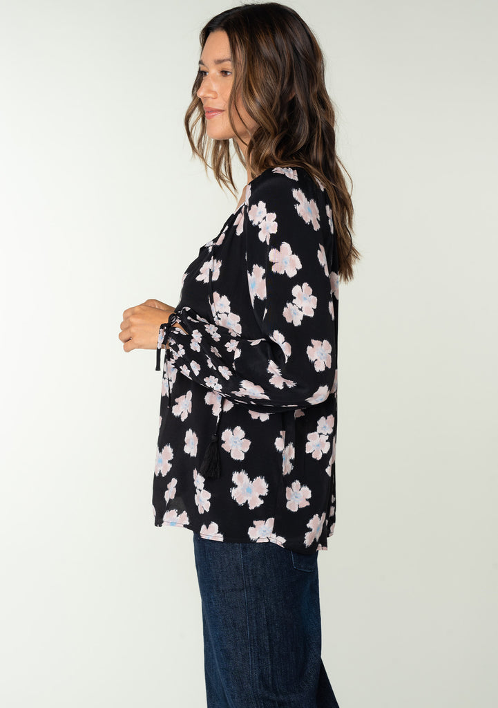 [Color: Black/Taupe] A side facing image of a brunette model wearing a flowy bohemian top in a black and taupe floral print. With long sleeves, adjustable tie wrist cuffs, and a split v neckline with tassel ties. 