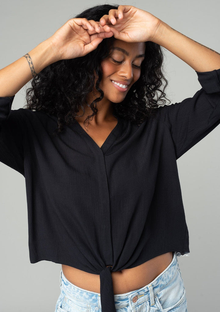 [Color: Black] A half body front facing image of a brunette model wearing a basic black blouse with long rolled sleeves, a button tab closure, a tie waist detail, and a concealed button front.  