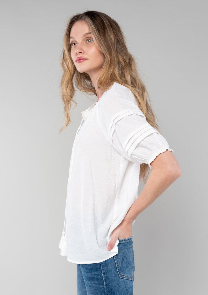 [Color: White] A side facing image of a blonde model wearing a classic bohemian white spring top with short puff sleeves, a button front, a split neckline with tassel ties, and delicate pintuck details. 