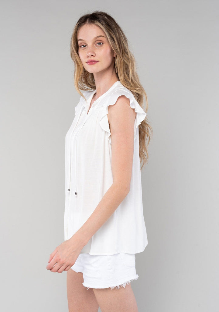 [Color: Vanilla] A side facing image of a blonde model wearing a solid white bohemian spring top. With short flutter cap sleeves, a self covered button front, neckline ties, and a relaxed fit. 