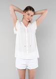 [Color: Vanilla] A front facing image of a blonde model wearing a solid white bohemian spring top. With short flutter cap sleeves, a self covered button front, neckline ties, and a relaxed fit. 