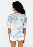 [Color: Ivory/Light Blue] A back facing image of a brunette model wearing a pretty spring top in a blue watercolor floral print. With short puff sleeves, a self covered loop button front, a v neckline, and a tie front waist. 