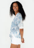 [Color: Ivory/Light Blue] A side facing image of a brunette model wearing a pretty spring top in a blue watercolor floral print. With short puff sleeves, a self covered loop button front, a v neckline, and a tie front waist. 