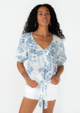 [Color: Ivory/Light Blue] A front facing image of a brunette model wearing a pretty spring top in a blue watercolor floral print. With short puff sleeves, a self covered loop button front, a v neckline, and a tie front waist. 