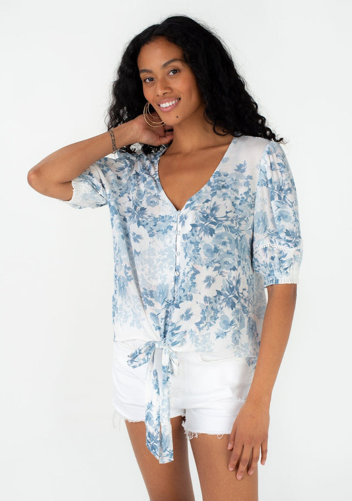 [Color: Ivory/Light Blue] A front facing image of a brunette model wearing a pretty spring top in a blue watercolor floral print. With short puff sleeves, a self covered loop button front, a v neckline, and a tie front waist. 