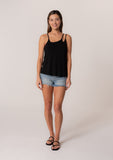 [Color: Black] A full body front facing image of a brunette model wearing a black stretchy bamboo knit tank top with asymmetric spaghetti straps, a scoop neckline, and a relaxed slim fit.