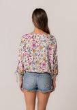 [Color: Natural/Yellow] A back facing image of a brunette model wearing a classic bohemian summer top in a mixed floral print. With three quarter length sleeves, a gathered sleeve detail with ties, a v neckline, a self covered button front, and an elastic waist. 