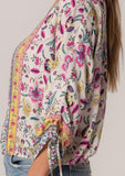 [Color: Natural/Yellow] A close up side facing image of a brunette model wearing a classic bohemian summer top in a mixed floral print. With three quarter length sleeves, a gathered sleeve detail with ties, a v neckline, a self covered button front, and an elastic waist. 