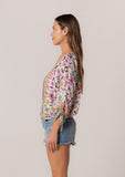 [Color: Natural/Yellow] A side facing image of a brunette model wearing a classic bohemian summer top in a mixed floral print. With three quarter length sleeves, a gathered sleeve detail with ties, a v neckline, a self covered button front, and an elastic waist. 