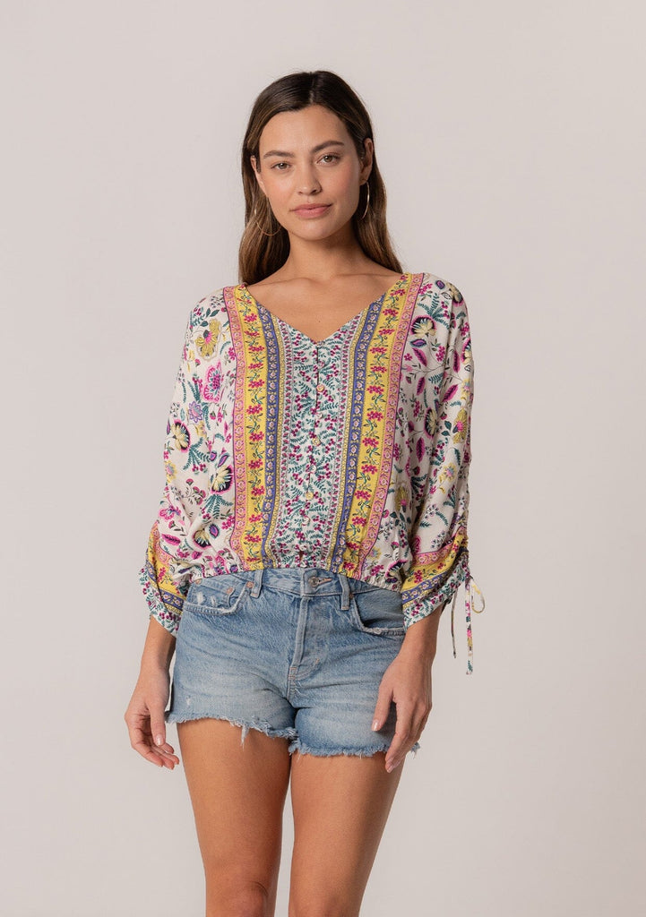 [Color: Natural/Yellow] A front facing image of a brunette model wearing a classic bohemian summer top in a mixed floral print. With three quarter length sleeves, a gathered sleeve detail with ties, a v neckline, a self covered button front, and an elastic waist. 