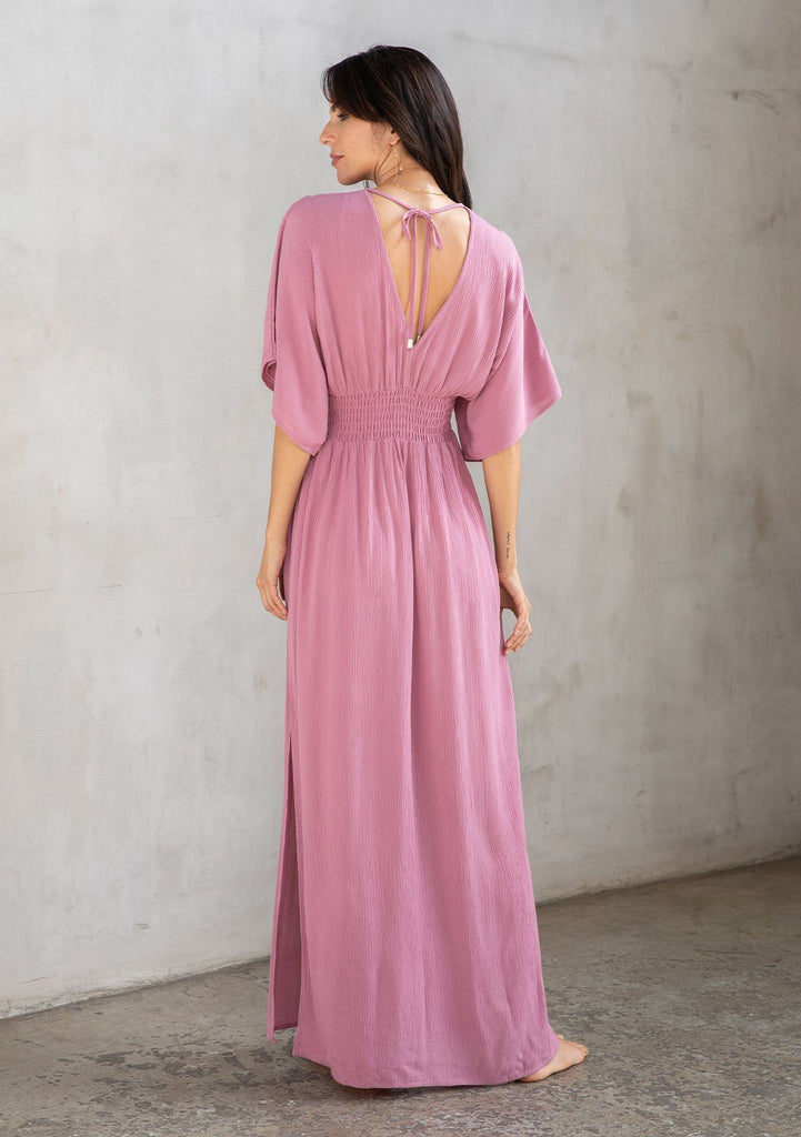 [Color: Smokey Orchid] A model wearing a resort ready light purple maxi dress. With half length kimono sleeves, a plunging v neckline, a smocked elastic empire waist, side slits, and an open back with tie closure.