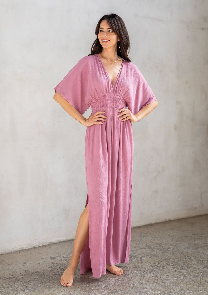 [Color: Smokey Orchid] A model wearing a resort ready light purple maxi dress. With half length kimono sleeves, a plunging v neckline, a smocked elastic empire waist, side slits, and an open back with tie closure.