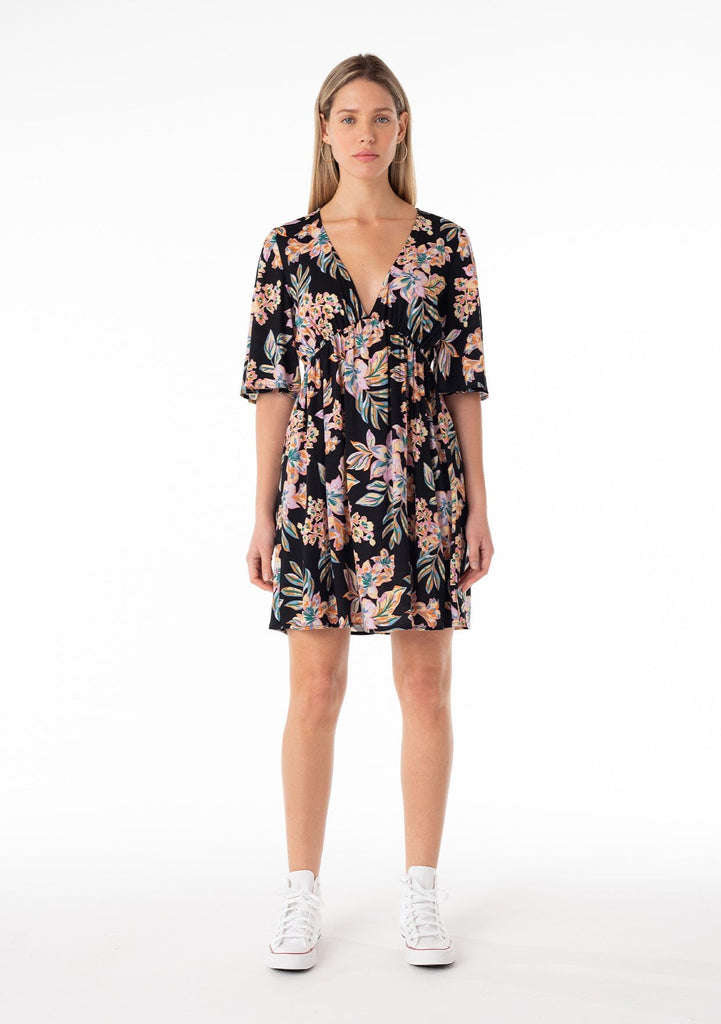 [Color: Black/Pink] A full body front facing image of a blonde model wearing a lightweight spring mini dress in a pink tropical floral print. With half length short sleeves, a deep v neckline, an empire waist, and a flowy, relaxed fit. 