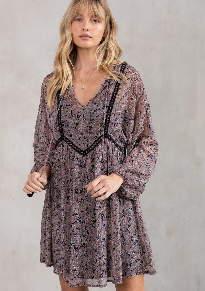 [Color: Black/Orchid] A model wearing a dreamy sheer chiffon bohemian black and purple paisley mini dress. With contrast crochet trim, a baby doll dress silhouette, and long sleeves. A flowy Fall bohemian mini dress.