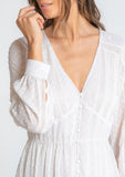 [Color: Gardenia] A model wearing a dreamy sheer off white maxi dress in swiss dot chiffon. With lace trim details throughout, a self covered button front, a sexy front slit, and sheer voluminous long sleeves with a button wrist cuff.