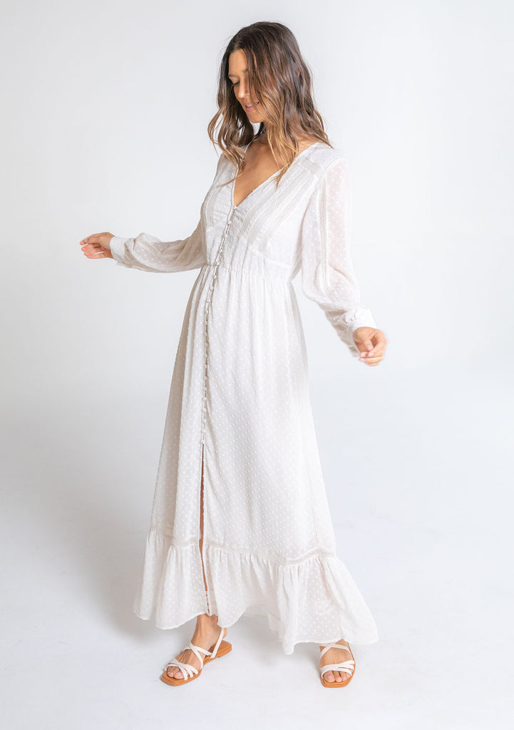 [Color: Gardenia] A model wearing a dreamy sheer off white maxi dress in swiss dot chiffon. With lace trim details throughout, a self covered button front, a sexy front slit, and sheer voluminous long sleeves with a button wrist cuff.