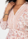 [Color: Natural/Clay] A close up front facing image of a brunette model wearing a bohemian spring mini dress in a pink floral border print. With voluminous three quarter length sleeves, a v neckline, a smocked elastic waist, a ruffle trimmed flowy tiered skirt, and an open back with tassel tie closure. 