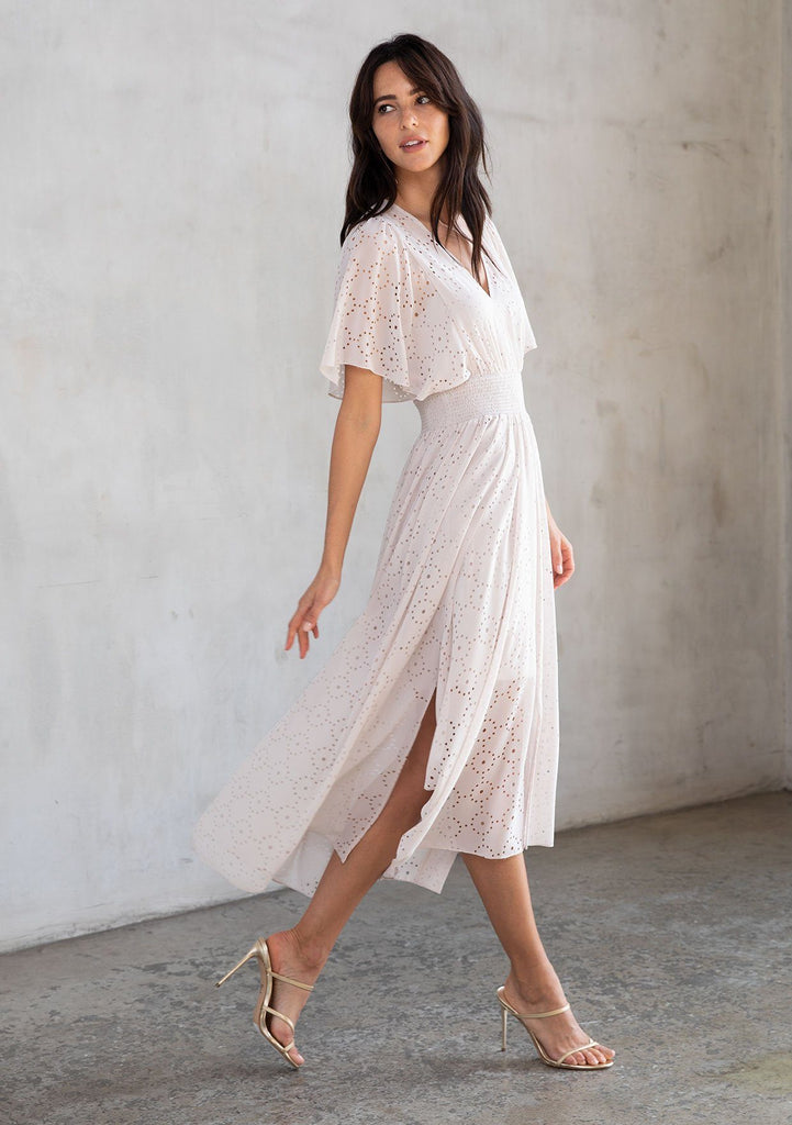 [Color: Bone] A model wearing a dreamy sheer off white chiffon maxi dress with gold trimmed laser cut details throughout. With a flowy skirt, short flutter sleeves, and a shark bite hemline.