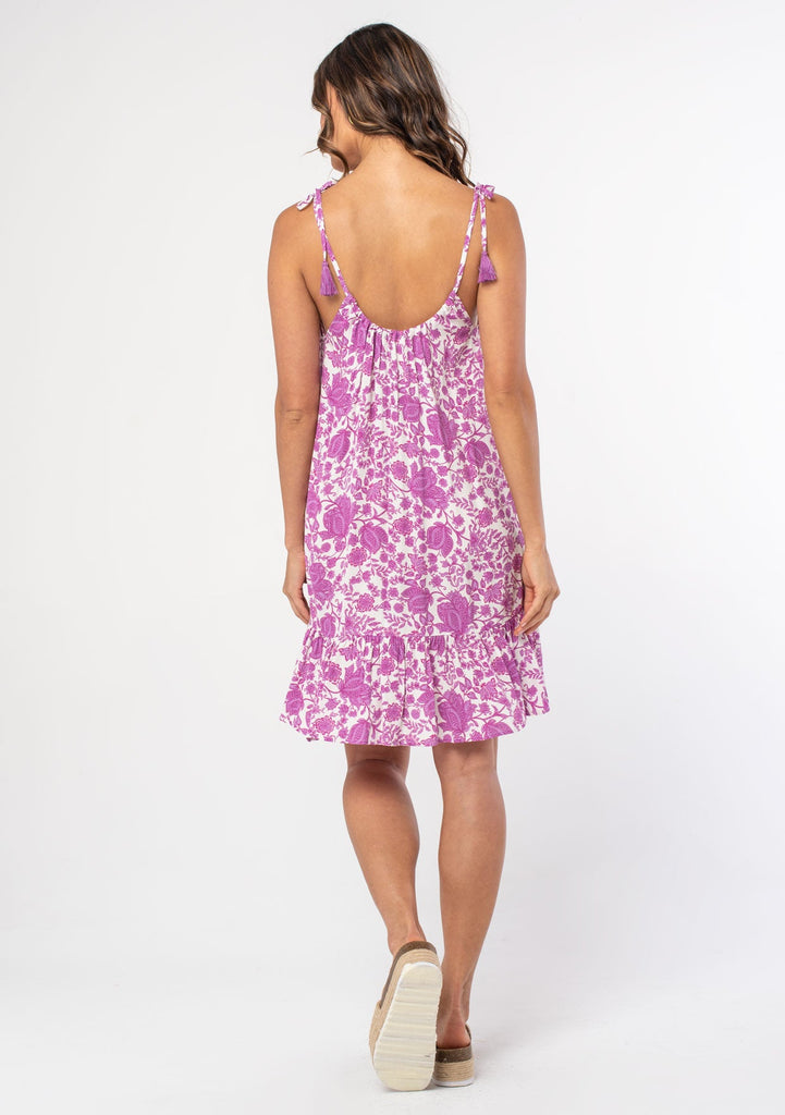 [Color: Ivory/Lilac] A model wearing a purple and white floral print mini tank dress with tie shoulder spaghetti straps, tassel ties, and a flowy fit.