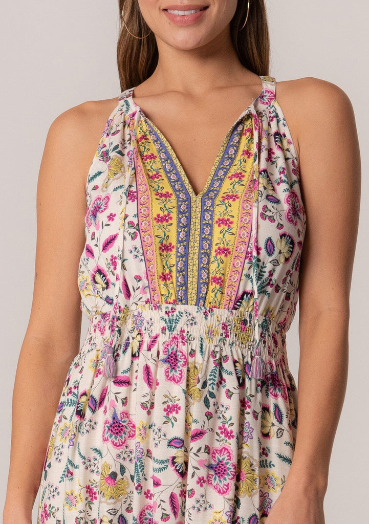 [Color: Natural/Yellow] A close up front facing image of a brunette model wearing a casual bohemian summer mini dress in a mixed floral print. A sleeveless dress with a racerback, a smocked elastic waist, a split v neckline with tassel ties, and a relaxed fit. 