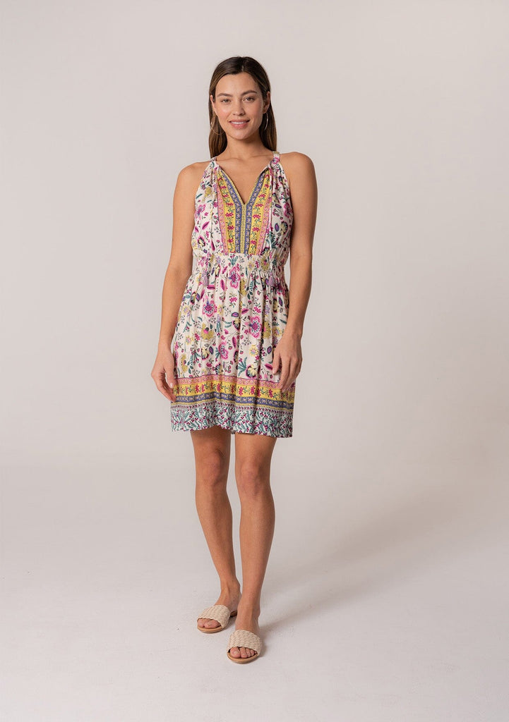 [Color: Natural/Yellow] A front facing image of a brunette model wearing a casual bohemian summer mini dress in a mixed floral print. A sleeveless dress with a racerback, a smocked elastic waist, a split v neckline with tassel ties, and a relaxed fit. 