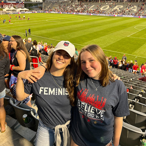 Woman on left wearing a white US Soccer hat and navy shirt that says "feminist". Woman on right wearing a navy shirt that says "I believe that we did win". They are inside a soccer stadium.