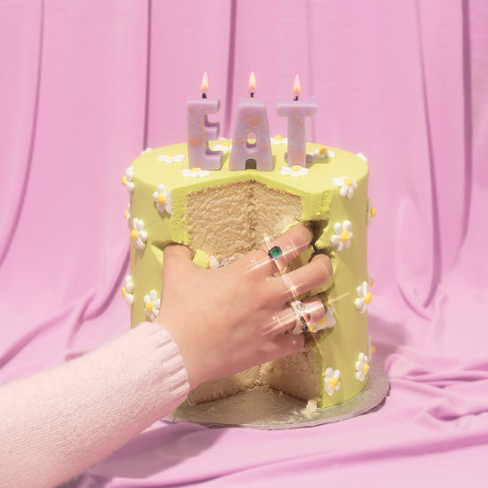 Woman's hand digging into a yellow cake that features daisies made of icing and lit candles that spell the word 