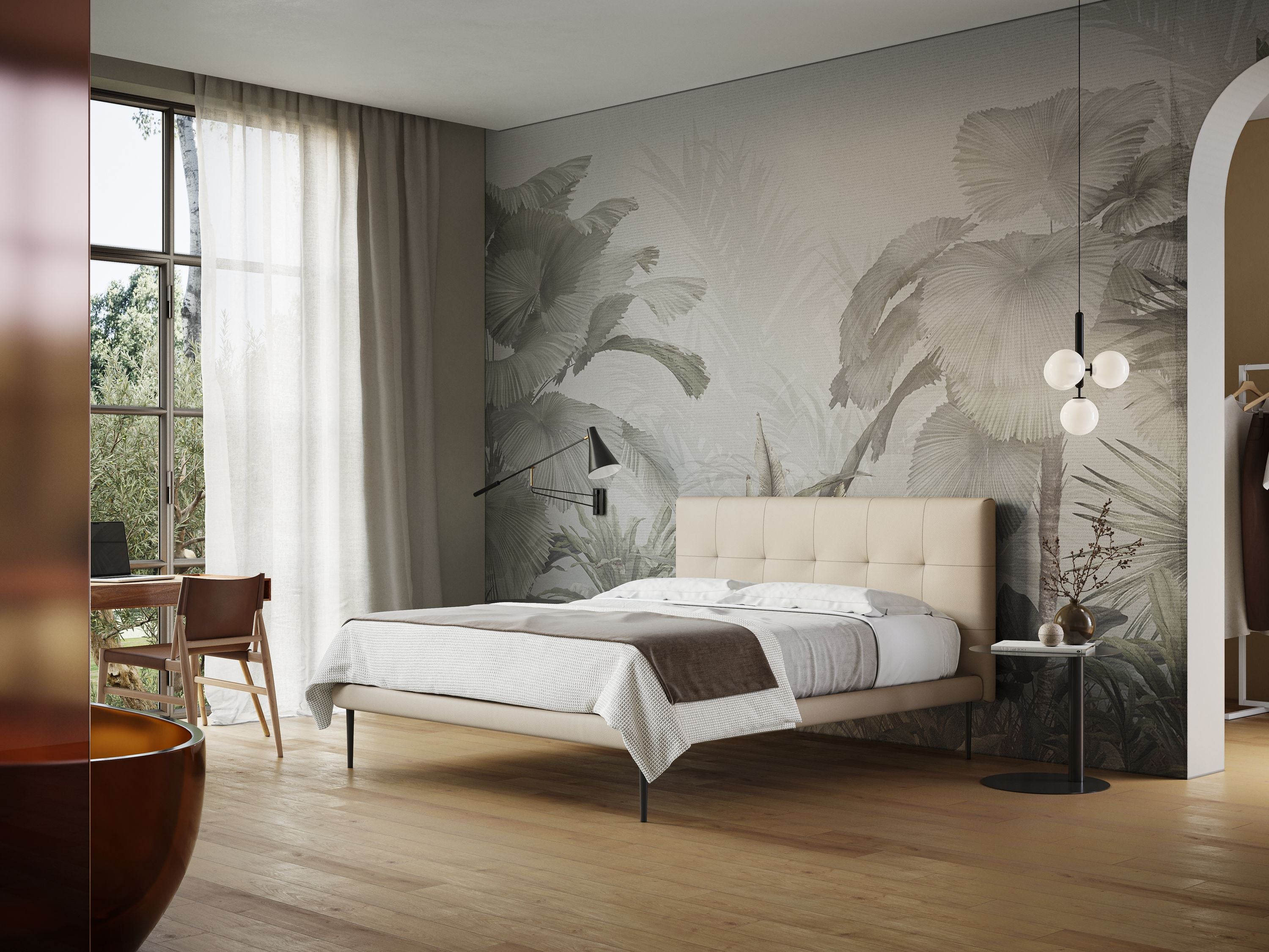 Una camera da letto moderna con un grande letto Midori imbottito trapuntato in ecopelle con piedini alti T04 di Letti.store, con testiera in ecopelle beige e biancheria da letto bianca e grigia. La stanza è decorata con carta da parati a tema natura e comprende una lampada da terra, un comodino rotondo decorato con vari oggetti e una scrivania in legno abbinata a una sedia accanto a un'ampia finestra drappeggiata con tende trasparenti che lasciano filtrare la luce naturale.