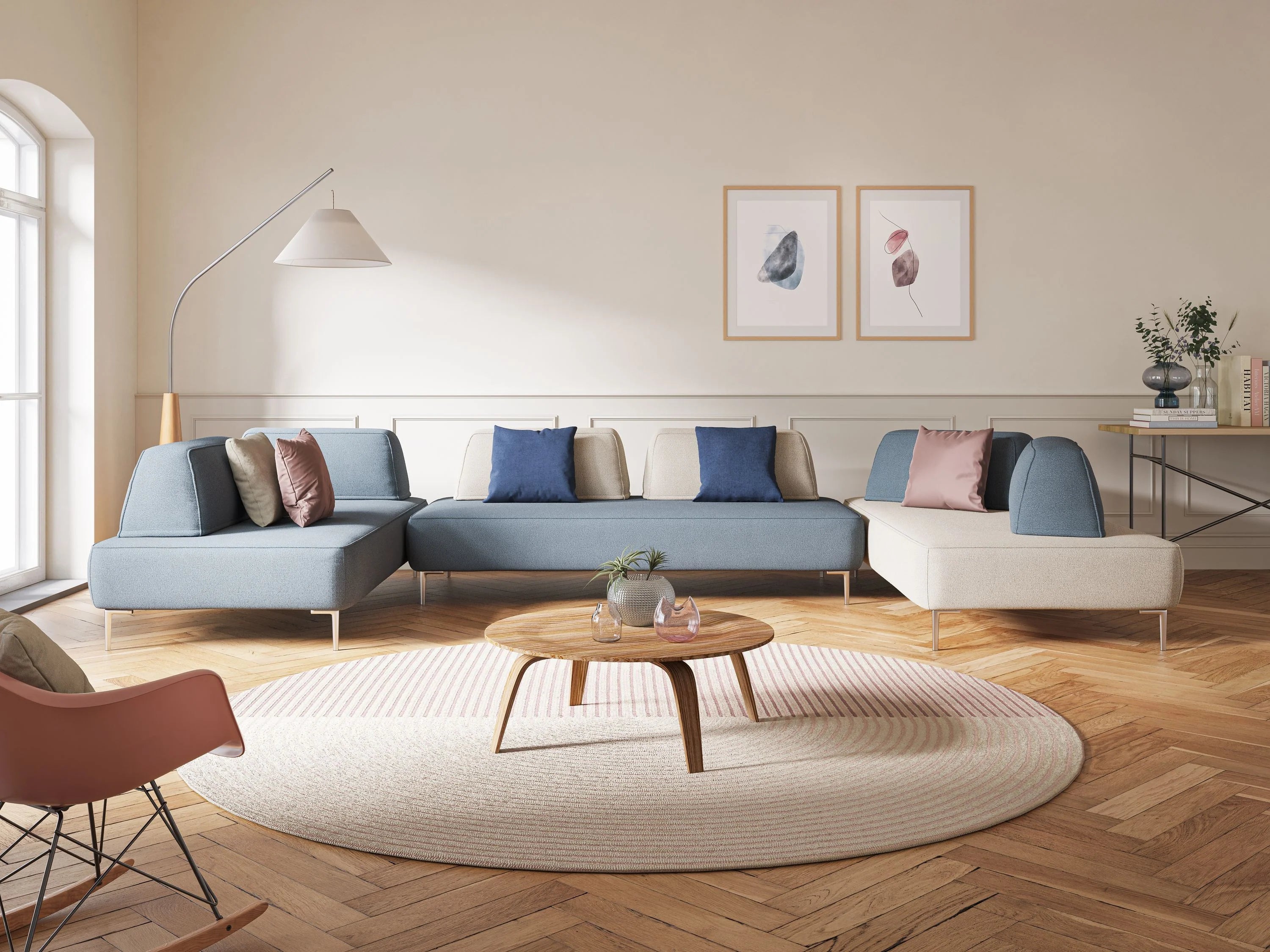 Un soggiorno moderno mette in mostra il divano panoramico modulare con schienali movibili T07 antracite di Divani.Store, cuscini blu e beige, un tavolino da caffè rotondo in legno su un tappeto circolare, quadri incorniciati su pareti color crema e finestre illuminate dal sole con tende trasparenti.