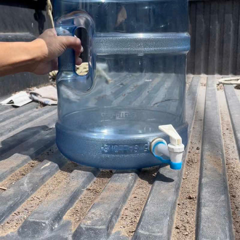a 5 gallon water cooler jug with a white dispenser tip covered be a blue cap to prevent dust from entering on a truck bed being held by a hand